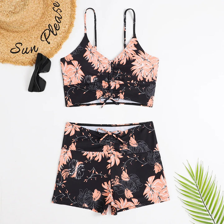 Dames zomerset met bloemenprint: zwarte crop top en shorts, strandhoed met tekst "Sun Please", zonnebril en palmblad. Perfect voor strandmode.